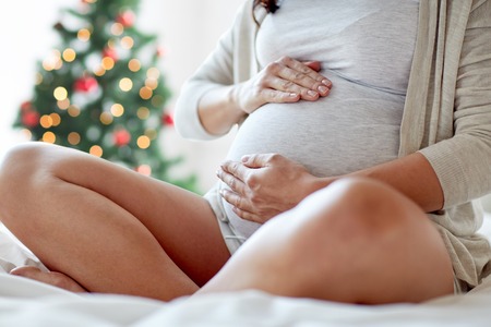 pregnancy, holidays, people and expectation concept - close up of pregnant woman sitting in bed and touching her belly at home over christmas tree backgroundの写真素材