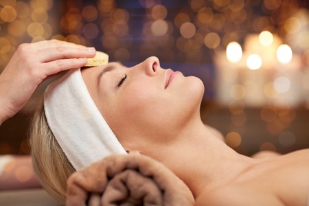 people, beauty, spa, cosmetology and relaxation concept - close up of beautiful young woman lying with closed eyes having face cleaning by sponge in spaの写真素材