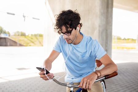 people, communication, technology, leisure and lifestyle - hipster man with smartphone and earphones on fixed gear bike listening to music on city streetの写真素材