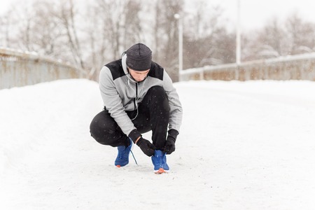 fitness, sport, people, music and healthy lifestyle concept - young man man in earphones tying shoe on winter bridgeの写真素材