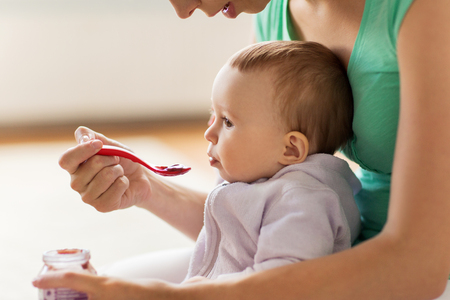 family, food, child, eating and parenthood concept - mother with puree and spoon feeding little baby at homeの写真素材