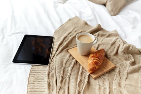 technology, coziness, morning and winter concept - tablet pc computer, coffee cup and croissant on bed at homeの写真素材