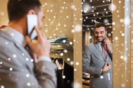 sale, shopping, fashion, style and people concept - happy young man or businessman trying suit and tie on and calling on smartphone at clothing store mirror over snowの写真素材