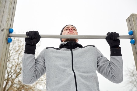 fitness, sport, exercising, training and people concept - young man doing pull ups on horizontal bar...