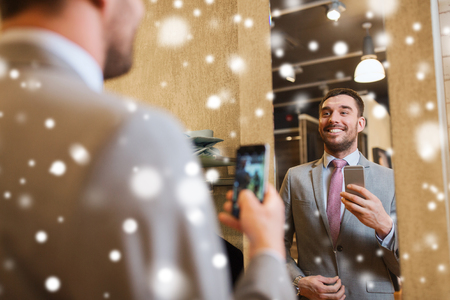 sale, shopping, fashion, style and people concept - happy young man or businessman in suit with smartphone taking mirror selfie at clothing store over snowの写真素材