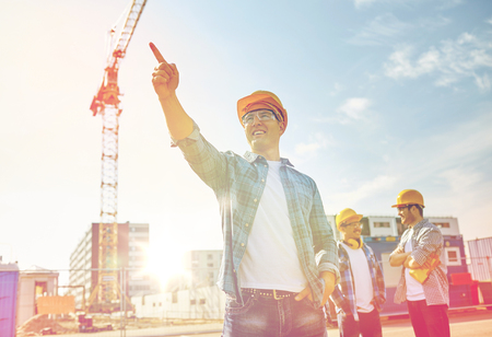 business, building, teamwork and people concept - group of smiling builders in hardhats pointing finger aside on construction siteの写真素材