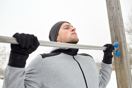 fitness, sport, exercising, training and people concept - young man doing pull ups on horizontal bar outdoors in winterの写真素材