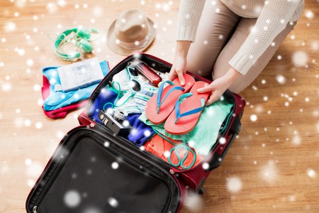 winter holidays, vacation, tourism and people concept - close up of woman packing bag with summer clothes and travel stuff over snowの写真素材
