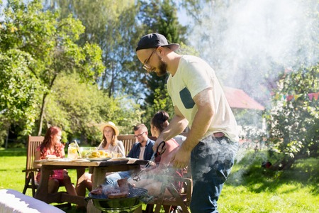 leisure, food, people and holidays concept - man cooking meat on barbecue grill for his friends at summer outdoor partyの写真素材