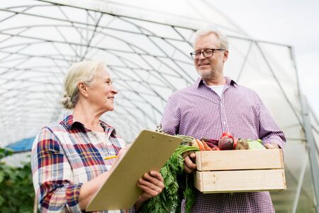 farming, gardening, agriculture, harvesting and people concept - senior couple with box of vegetables and clipboard at farm greenhouseの写真素材