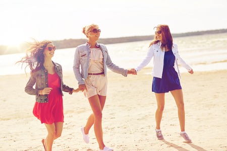summer vacation, holidays, travel and people concept - group of smiling young women in sunglasses and casual clothes running along beachの写真素材