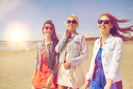 summer vacation, holidays, travel and people concept - group of smiling young women in sunglasses and casual clothes walking along beachの写真素材