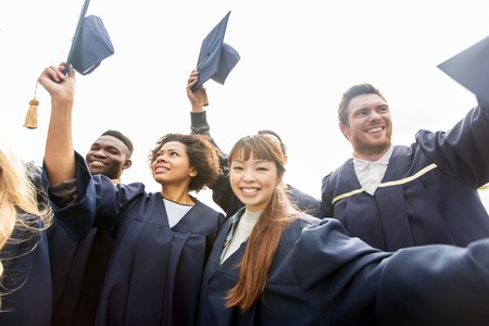 education, graduation and people concept - group of happy international students in bachelor gowns waving mortar boards or hatsの写真素材