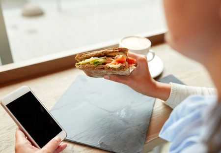 food, dinner, technology and people concept - woman with smartphone eating salmon panini sandwich with tomatoes and cheese at restaurantの写真素材