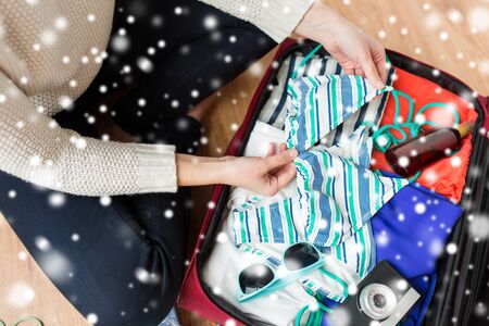 winter holidays, vacation, tourism and objects concept - close up of woman packing travel bag with bikini, camera and summer clothes over snowの写真素材