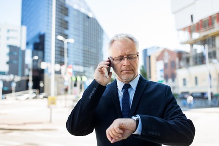 business, technology, time, punctuality and people concept - senior businessman calling on smartphone an looking at wristwatch or smart watch on his hand in cityの写真素材