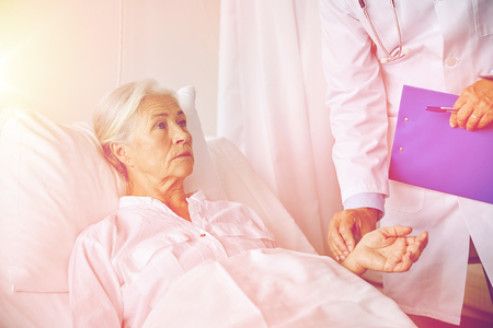 medicine, age, health care and people concept - doctor with clipboard visiting senior patient woman and checking her pulse at hospital wardの写真素材
