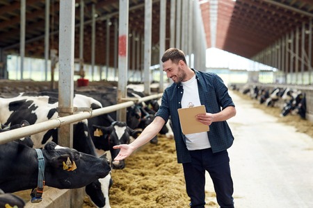 agriculture industry, farming, people and animal husbandry concept - happy smiling young man or farmer with clipboard and cows in cowshed on dairy farm agriculture industry, farming, people and animal husbandry concept - happy smiling young man or farmer with clipboard and cows in cowshed on dairy farm