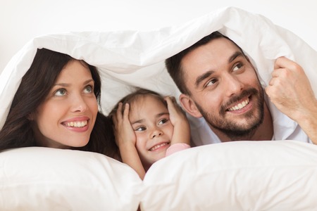 happy family lying in bed under blanket at homeの写真素材