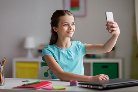 people, children and technology concept - girl with laptop computer and smartphone taking selfie at homeの写真素材