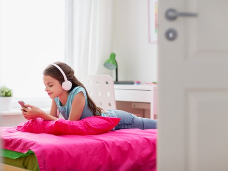 people, children, technology and leisure concept - happy girl with headphones and smartphone lying on bed and listening to music at homeの写真素材