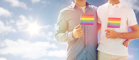 people, homosexuality, same-sex marriage and love concept - close up of happy male gay couple hugging and holding rainbow flags over sky and sun backgroundの写真素材
