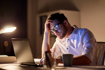 business, overwork, people, deadline and technology concept - stressed businessman in glasses with laptop computer thinking at night officeの写真素材