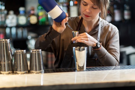 drinks, people and luxury concept - barmaid with shaker pouring alcohol from bottle into jigger and preparing cocktail at barの写真素材
