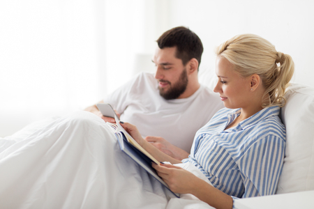 couple with book and smartphone in bed at homeの写真素材
