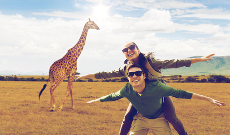 travel, tourism and people concept - happy couple with backpacks having fun over giraffe in african savannah backgroundの写真素材