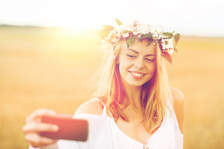 technology, summer holidays, vacation and people concept - smiling young woman in wreath of flowers taking selfie by smartphone on cereal fieldの写真素材