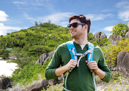 travel, tourism and people concept - happy young man in sunglasses with backpack over natural exotic island backgroundの写真素材