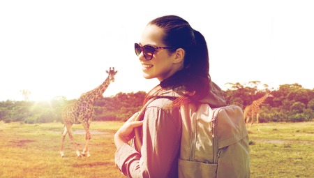 travel, tourism and people concept - happy young woman with backpack over african savannah and giraffe backgroundの写真素材