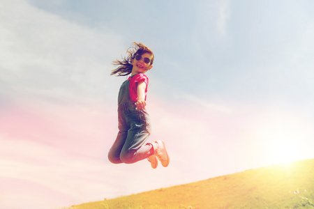 summer, childhood, leisure and people concept - happy little girl jumping high over green field and blue sky outdoorsの写真素材