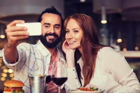 leisure, technology, date, people and holidays concept - happy couple having dinner and taking selfie by smartphone at restaurantの写真素材