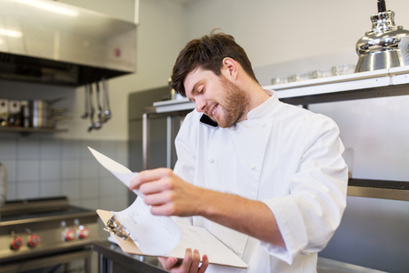 cooking, profession and people concept - male chef cook with clipboard calling on smartphone at restaurant kitchenの写真素材