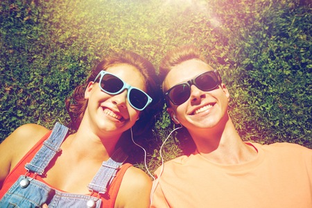 love and people concept - happy teenage couple in sunglasses lying on grass with earphones and listening to music at summerの写真素材