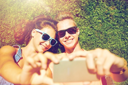 love and people concept - happy teenage couple in sunglasses lying on grass and taking selfie on...