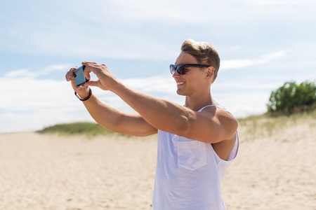 summer holidays and people concept - happy smiling young man with smartphone on beach photographingの写真素材