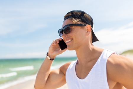 summer holidays and people concept - happy smiling young man in sunglasses calling on smartphone on beachの写真素材