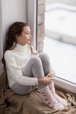 sad girl sitting on sill at home window in winterの写真素材