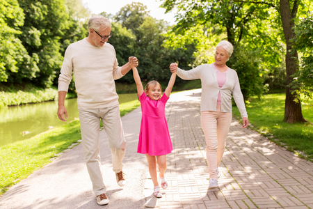 family, generation and people concept - happy smiling grandmother, grandfather and little granddaughter walking at parkの写真素材