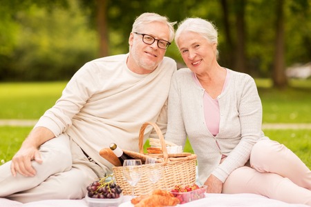 old age, holidays, leisure and people concept - happy senior couple with picnic basket sitting on blanket at summer parkの写真素材