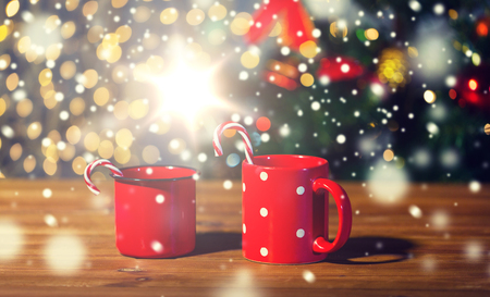 holidays, christmas, winter, food and drinks concept - close up of candy canes and cups on wooden table over lightsの写真素材