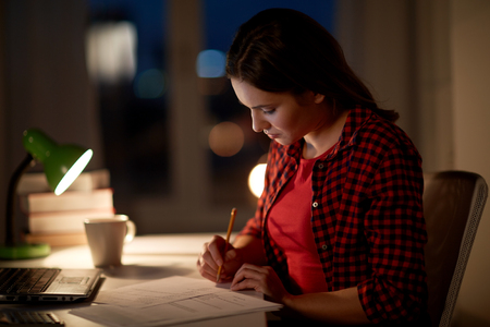 people, education, freelance and learning concept - woman or student girl with laptop computer writing to notebook at home at nightの写真素材