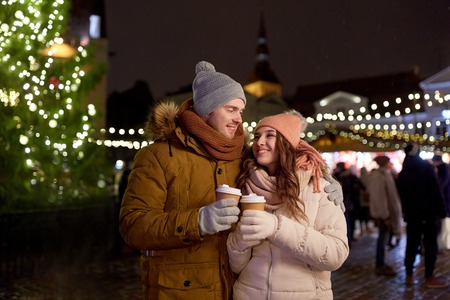 winter holidays, hot drinks and people concept - happy young couple with coffee at christmas market in eveningの写真素材