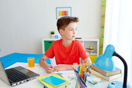 education, childhood and people concept - happy student boy with laptop computer writing to notebook...