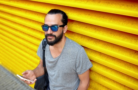 technology, lifestyle and people concept - man in sunglasses with bag and smartphone on street over ribbed yellow wall backgroundの写真素材