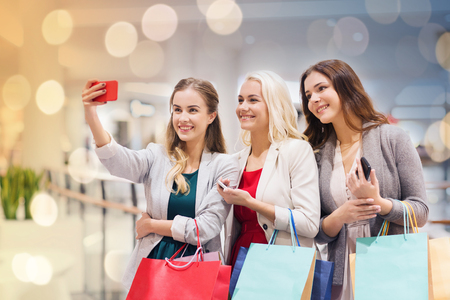 sale, consumerism, technology and people concept - happy young women with smartphones and shopping bags taking selfie in mallの写真素材