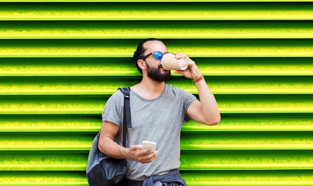 drinks, technology and people concept - man with smartphone drinking coffee from disposable paper cup on street over ribbed green wall backgroundの写真素材